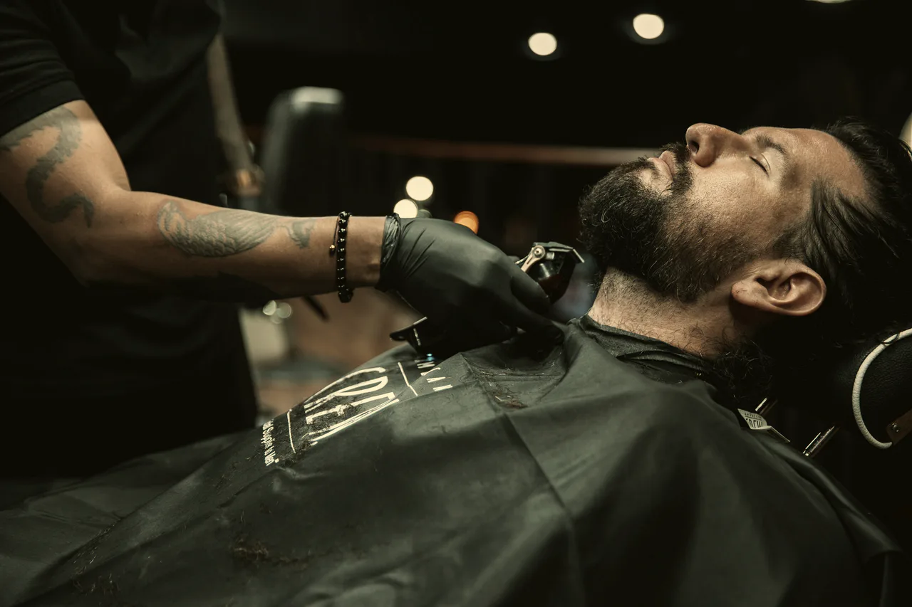 A barber in black gloves trims a reclined client's beard under warm shop lights